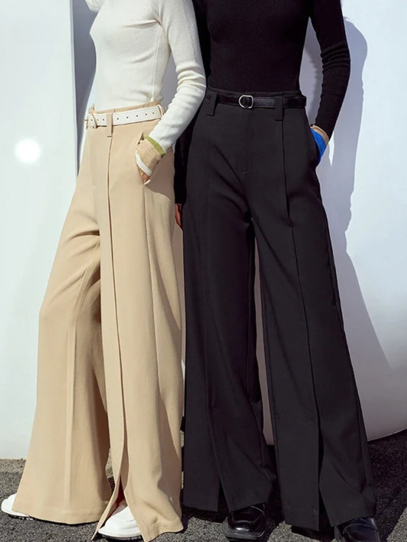 Two women wearing tailored, pleated, high waisted pants with slits at the hem.