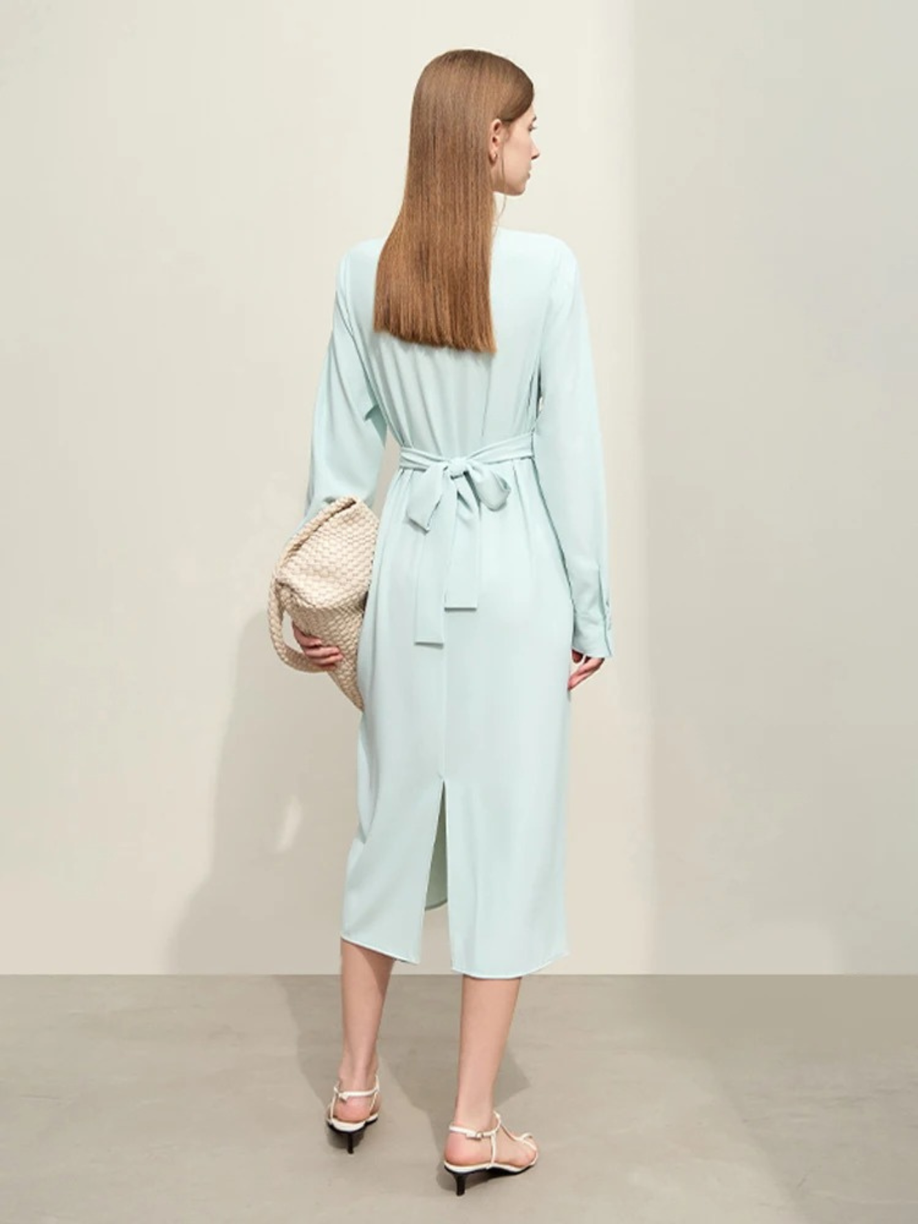 A woman showing the back of a blue, mid-calf length, long sleeved, flowy dress with a bow on the back to cinch the waist and a cut out at the hem for ease of wear.
