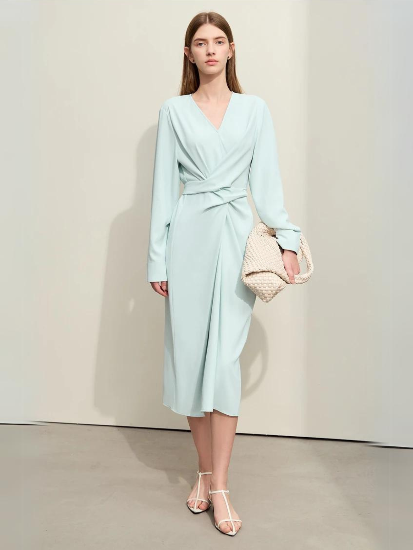 A woman wearing a light blue, mid-calf length, long sleeved dress with a v-neckline and a draping design on the bodice.