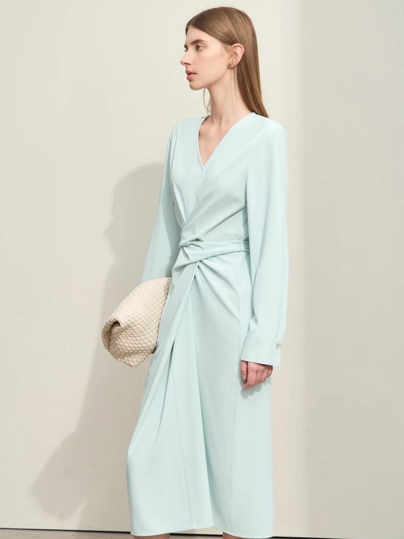 A woman wearing a light blue, mid-calf length, long sleeved dress with a v-neckline and a draping design on the bodice holding a white bag.