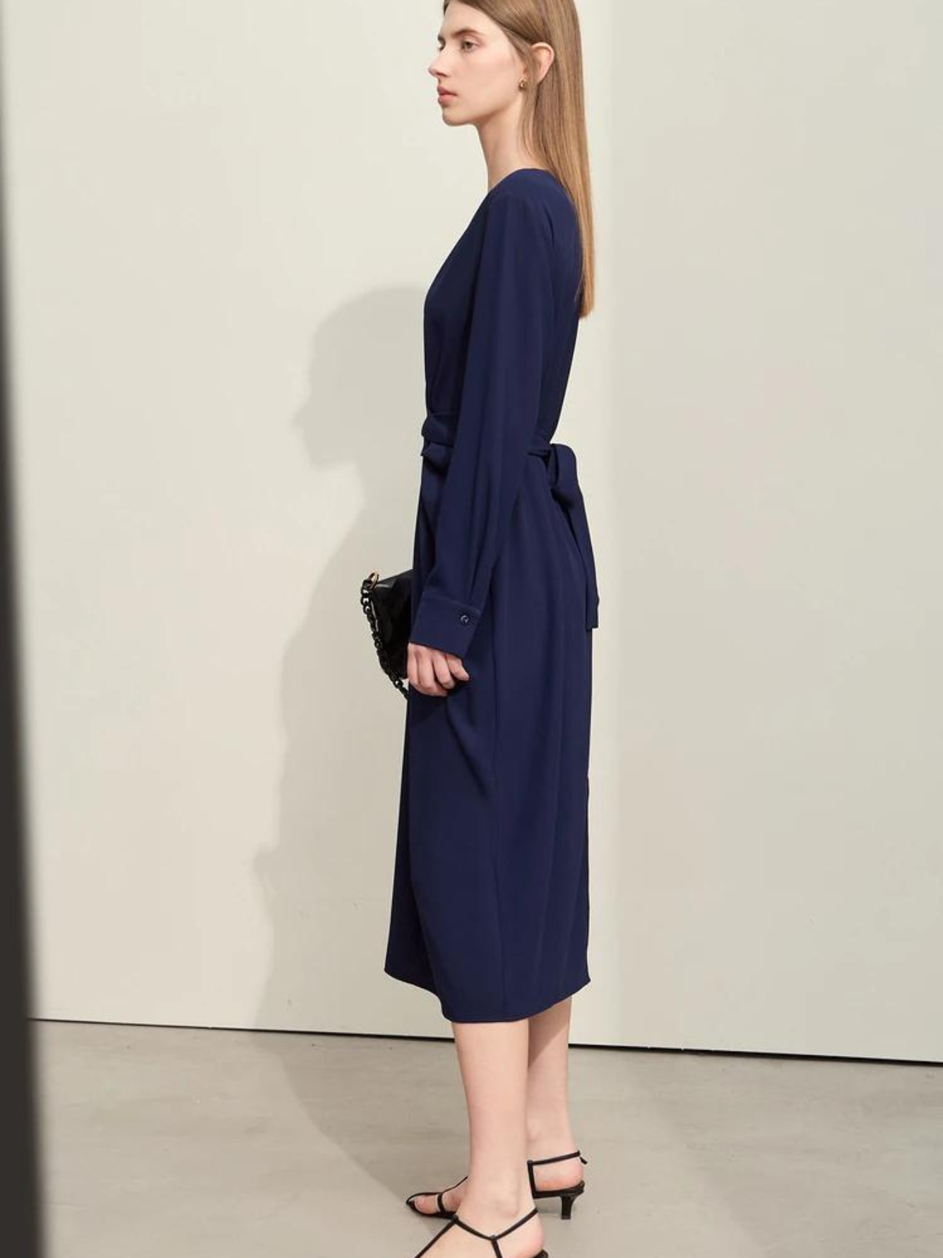 A woman wearing a dark blue, mid-calf length, long sleeved dress with a v-neckline and a draping design on the bodice and a bow on the back to cinch the waist.