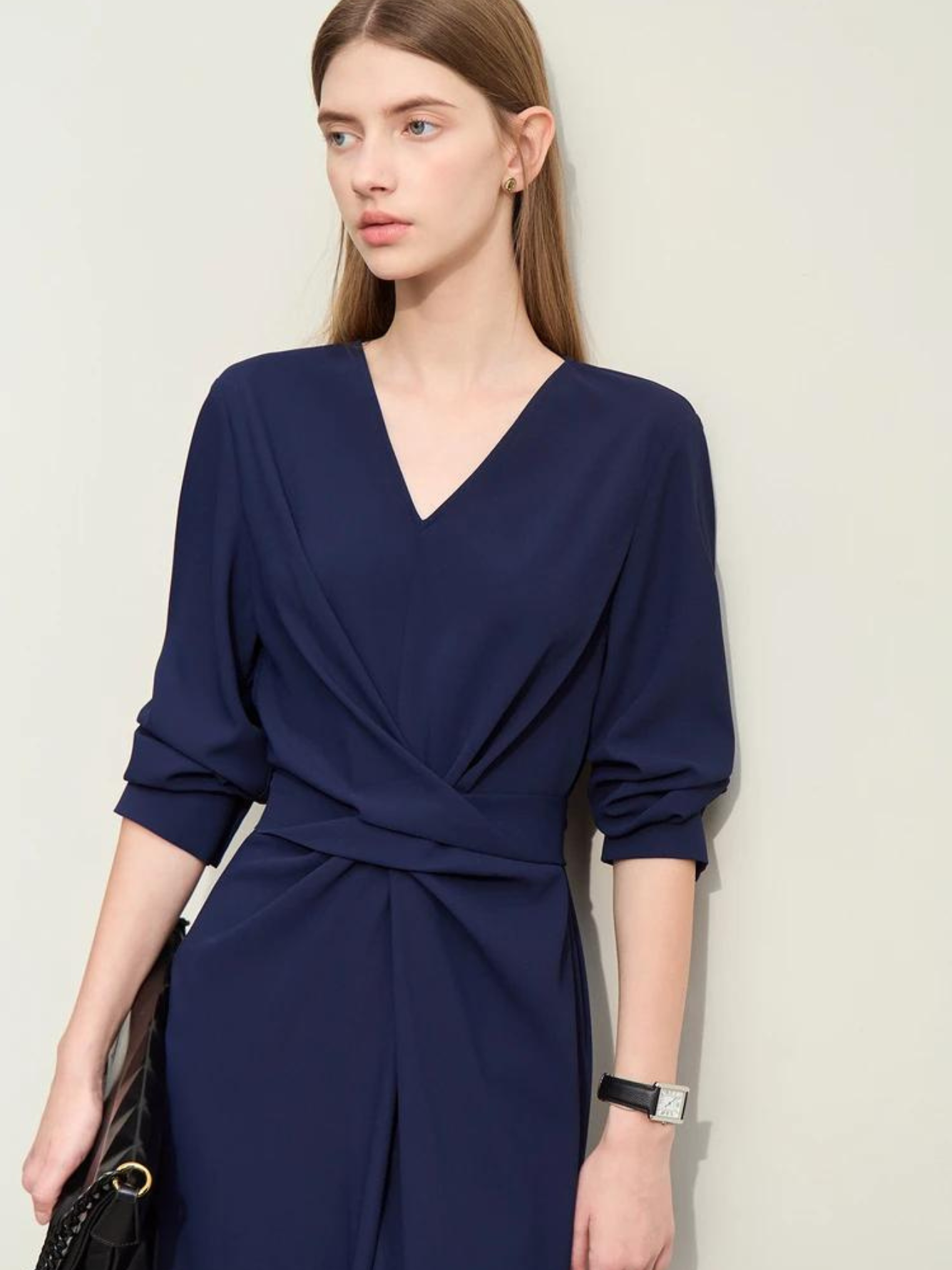 A woman wearing a dark blue, mid-calf length, long sleeved dress with a v-neckline and a draping design on the bodice and the sleeves rolled up for styling purposes.