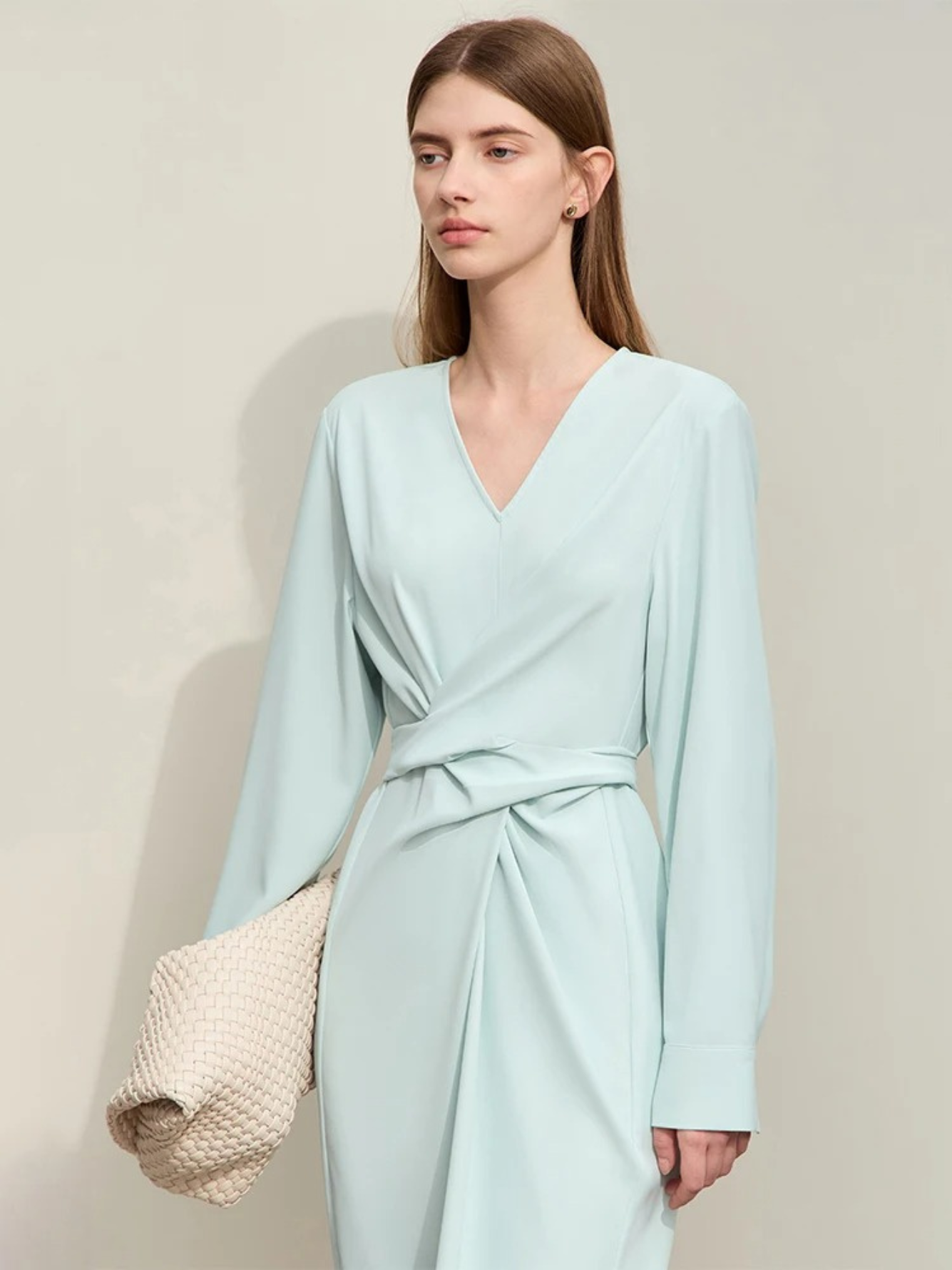 A woman wearing a light blue, mid-calf length, long sleeved, flowy dress with a v-neckline and a draping design on the bodice