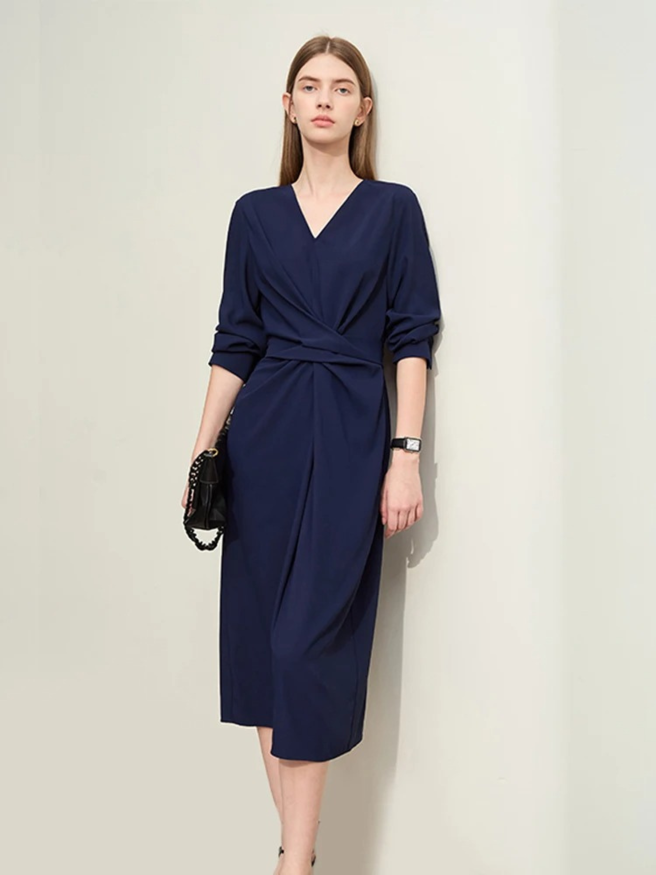 A woman wearing a dark blue, mid-calf length, long sleeved, flowy dress with a v-neckline and a draping design on the bodice with the sleeves rolled up for styling purposes.