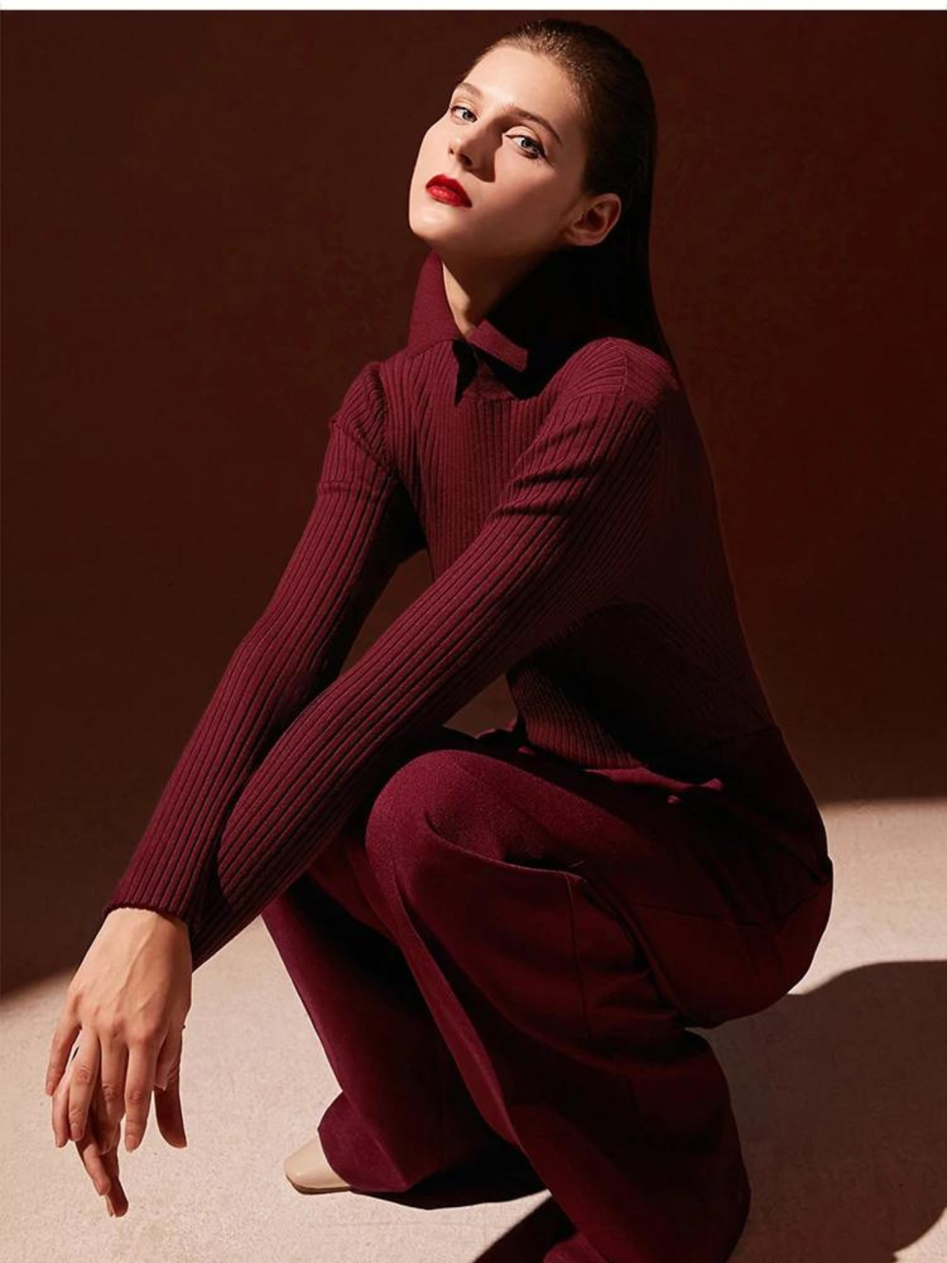 A brunette woman in a sitting position wearing a deep red sweater with a polo inspired collar and red tailored pants.