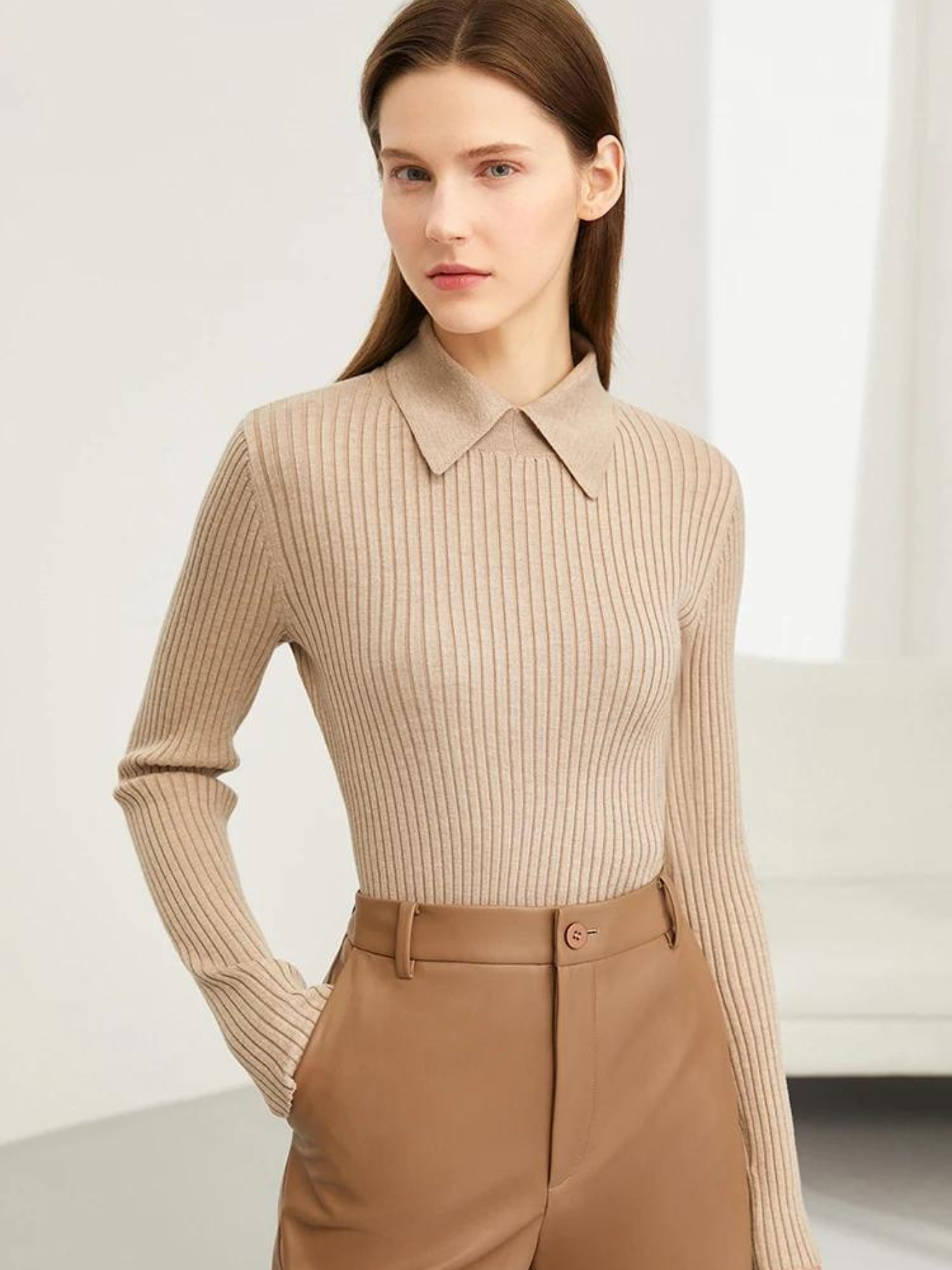 A brunette woman wearing a camel-colored sweater with a polo inspired collar and a pair of leather khaki shorts with one hand in her pocket.