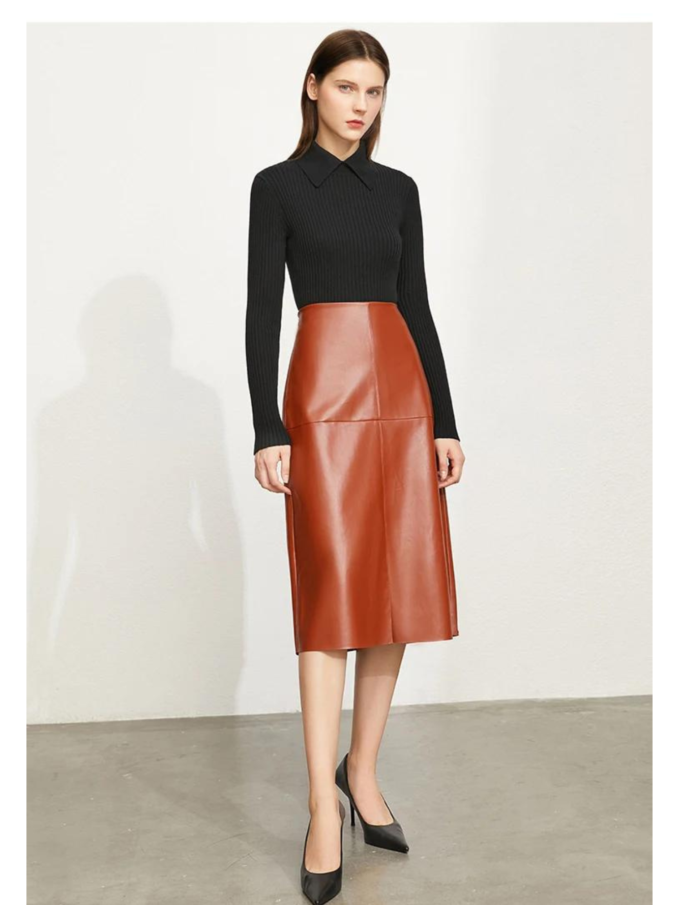 A brunette woman wearing a black fitted sweater with a polo inspired collar, a brown leather skirt, and a pair of black leather shoes..