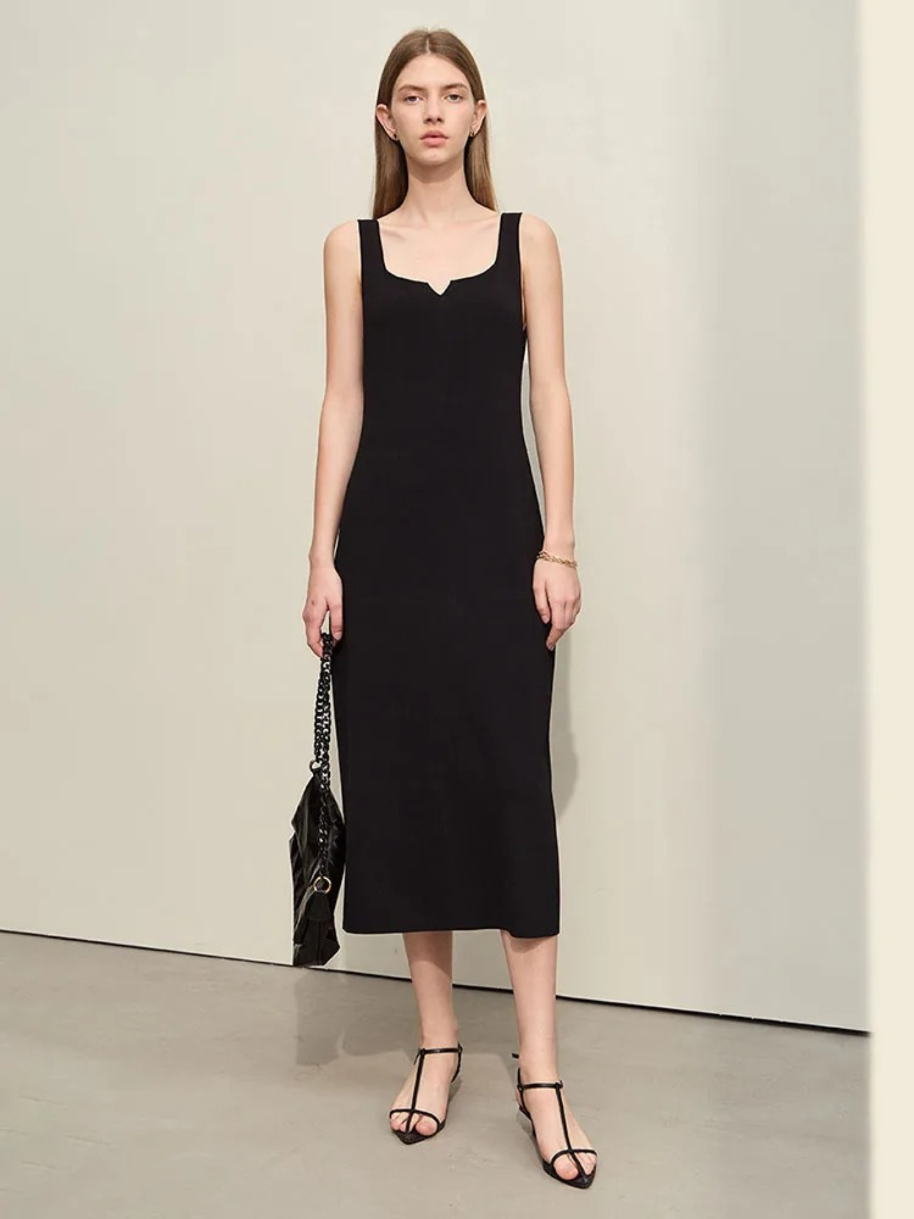 A woman wearing a minimalist, knee length, sleeveless dresses with thin straps and a mini cut out in the neckline in black paired with black strappy sandals and a bag.