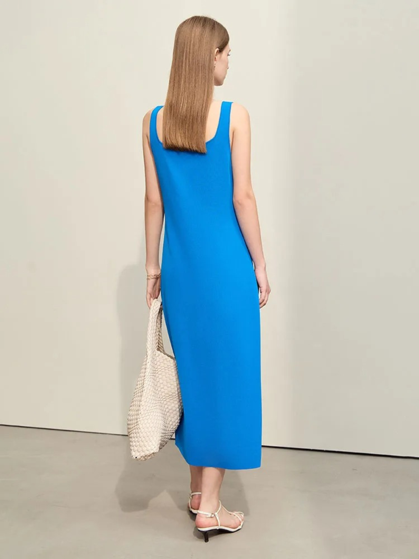 A woman showing the back of a minimalist, knee length, sleeveless dress with thin straps in blue paired with simple shoes and a bag.