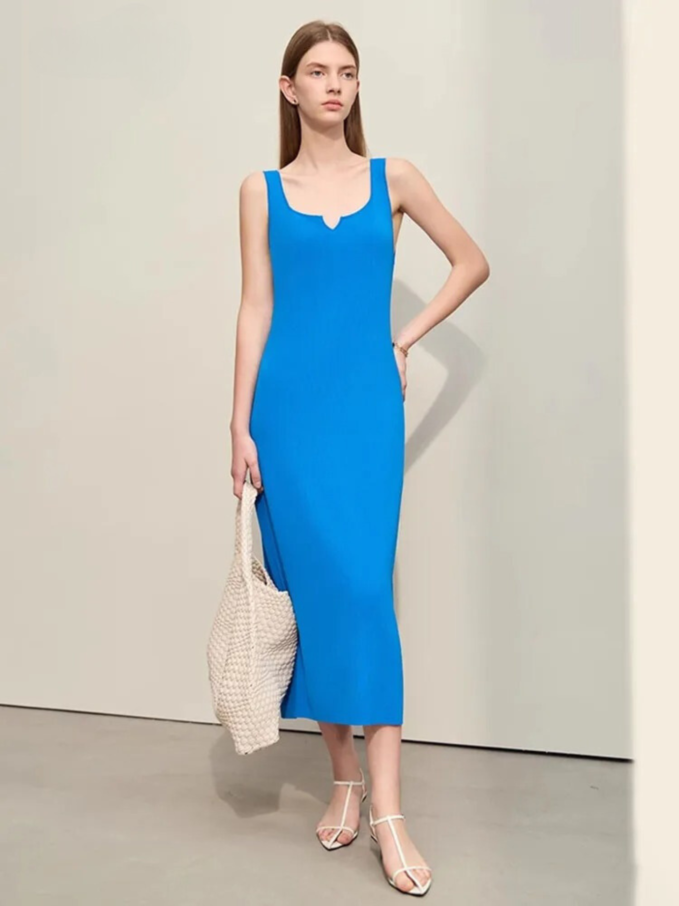 A woman wearing a minimalist, blue, mid-calf length dress with thin straps and a small cut out design on the neckline paired with simple open toed sandals and a bag.