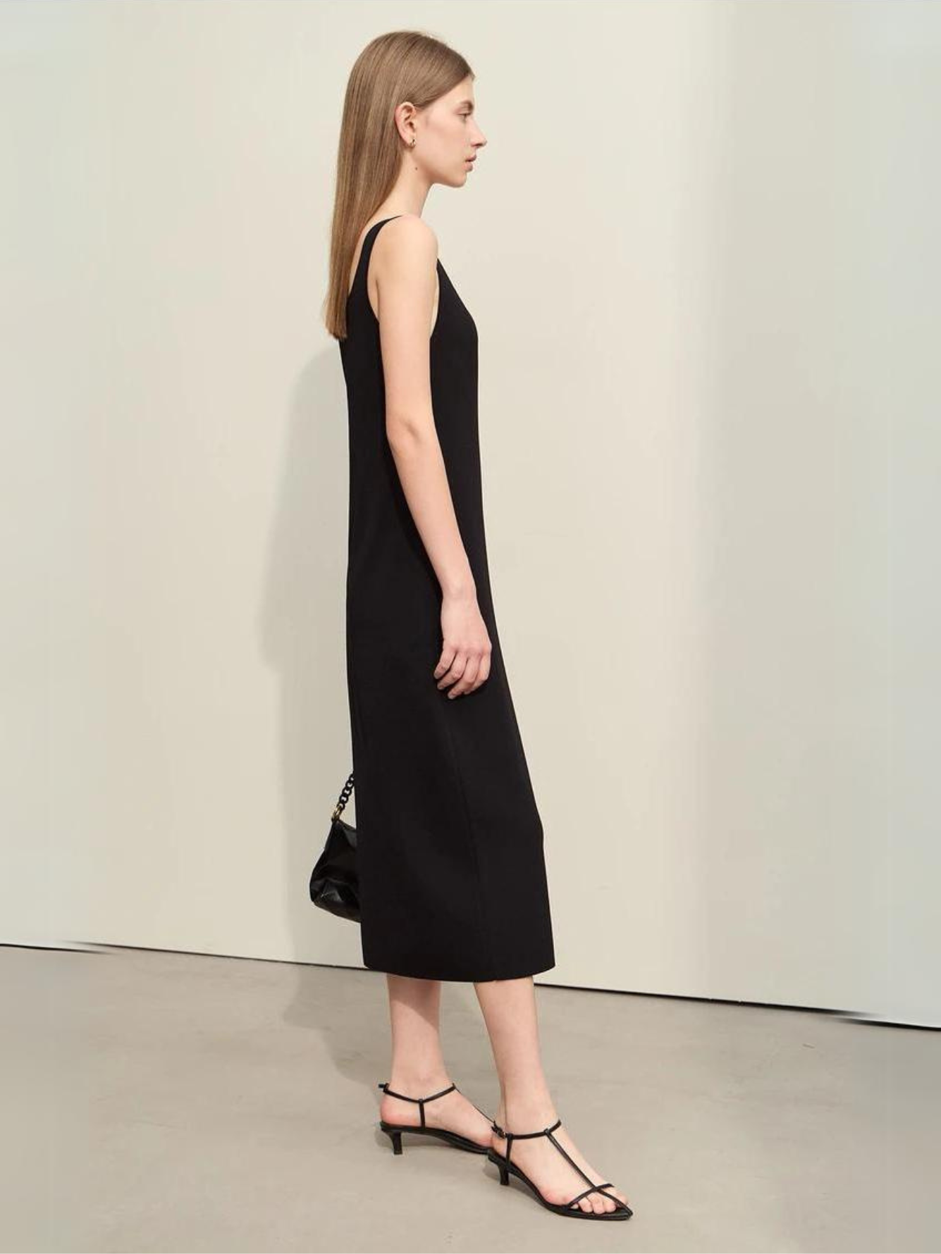 A woman wearing a minimalist, knee length, sleeveless dresses with thin straps and a mini cut out in the neckline in black.