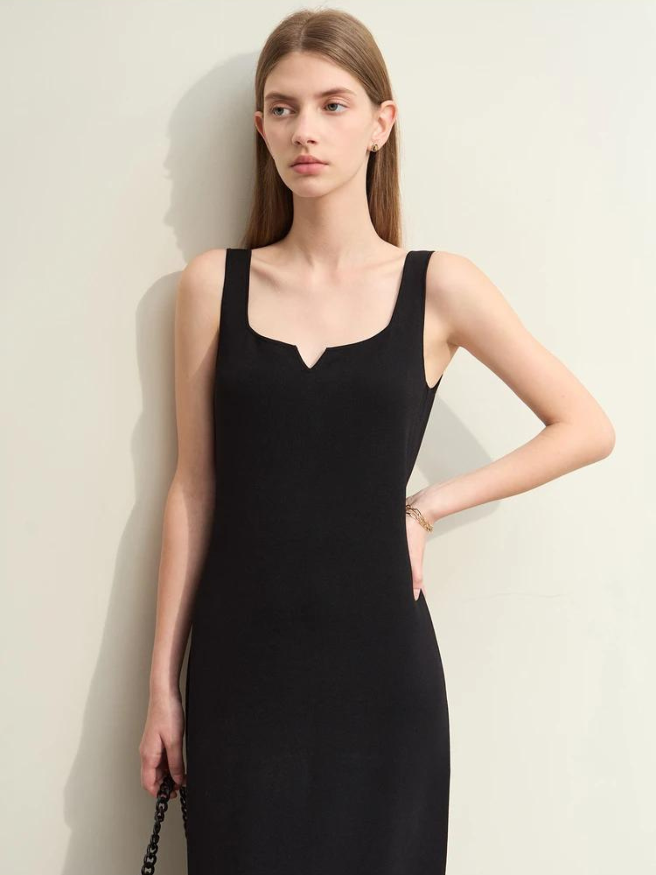 A woman wearing a minimalist, knee length, sleeveless dresses with thin straps and a mini cut out in the neckline in black.