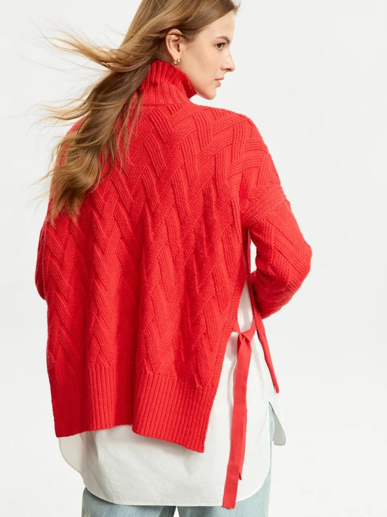 A woman with her back turned and wind blowing through her hair wearing a red knit sweater with a turtleneck neckline and tie design on the sides.