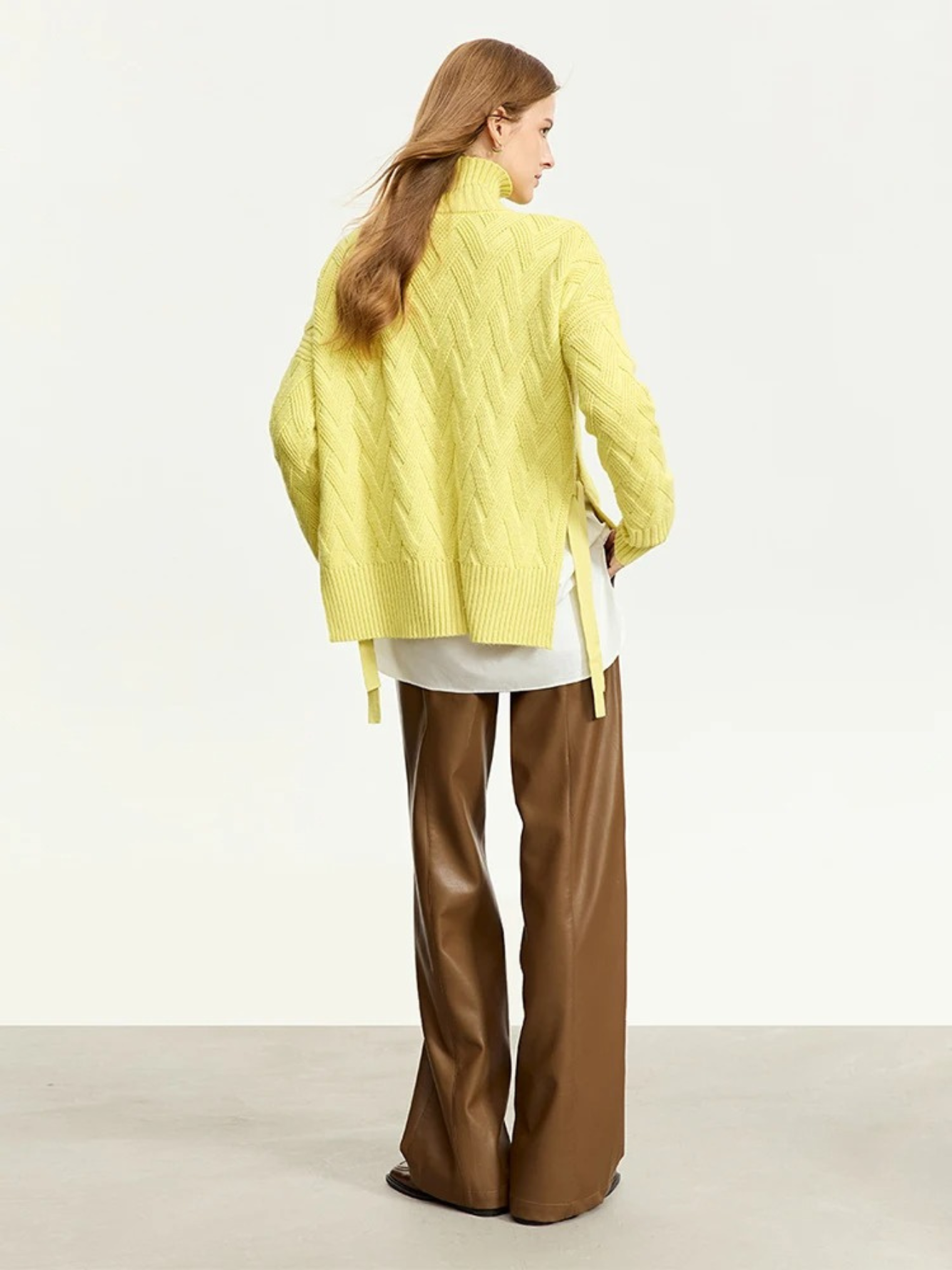 A woman wearing a mustard colored knit turtleneck sweater with a tie design on both sides over a white shirt and brown leather pants.