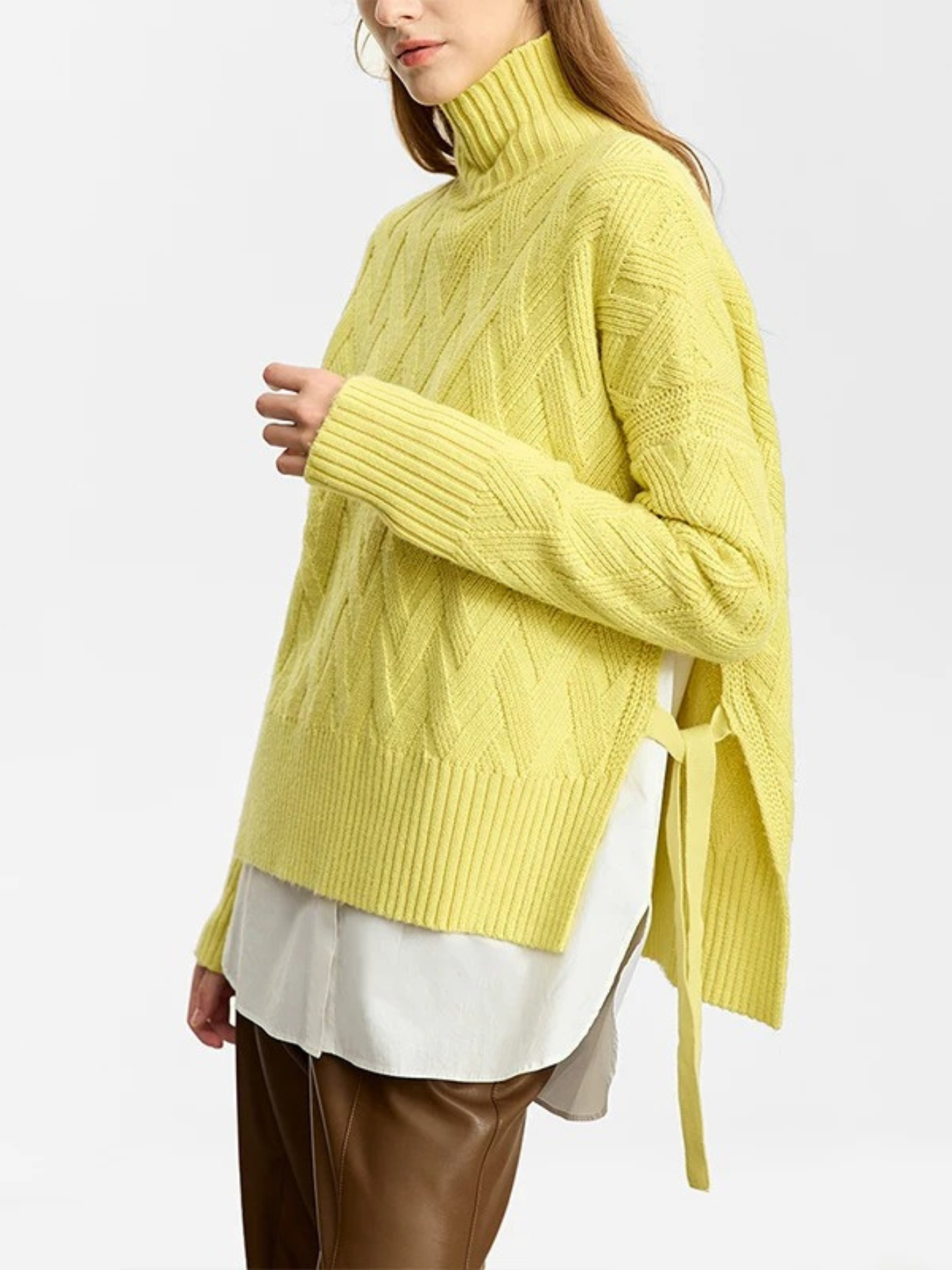 A woman wearing a mustard colored knit turtleneck sweater with a tie design on both sides over a white shirt and brown leather pants.