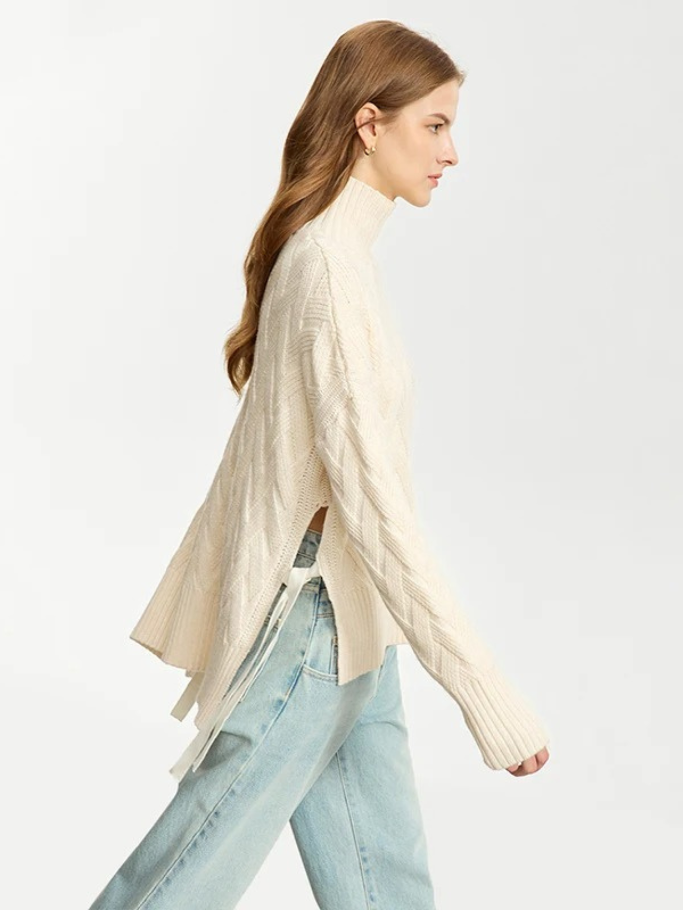 A woman wearing a cream colored knit turtleneck sweater with a tie design on both sides and a chevron knitting pattern and a pair of light denim jeans.
