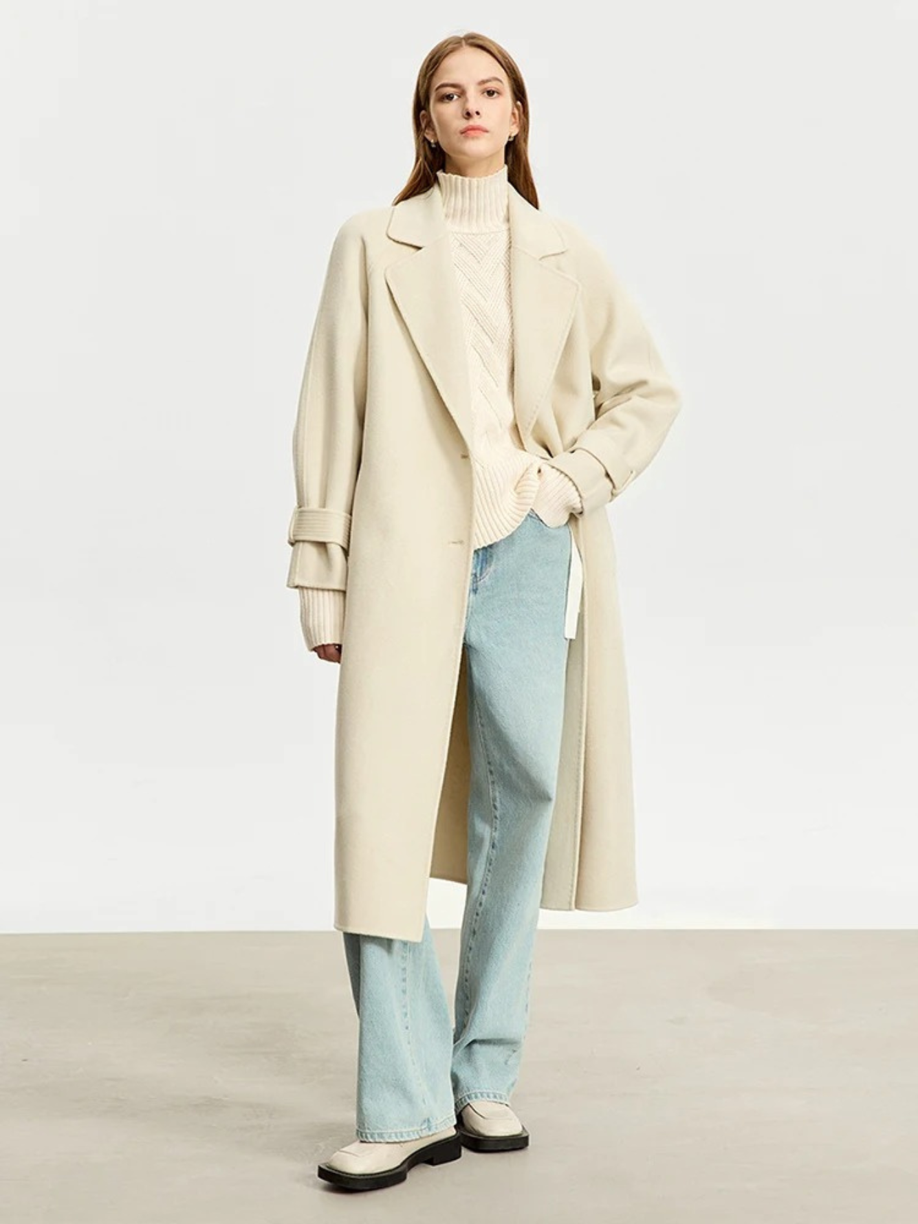A woman wearing a cream colored knit turtleneck sweater with a tie design on both sides and a chevron knitting pattern, a cream colored oversized coat, and pair of light washed denim jeans.