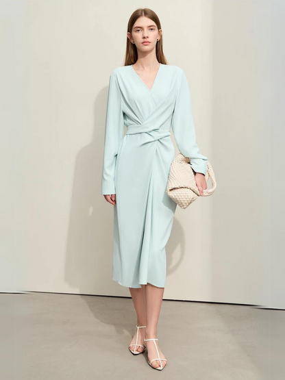 A woman wearing a light blue, mid-calf length, long sleeved dress with a v-neckline and a draping design on the bodice.