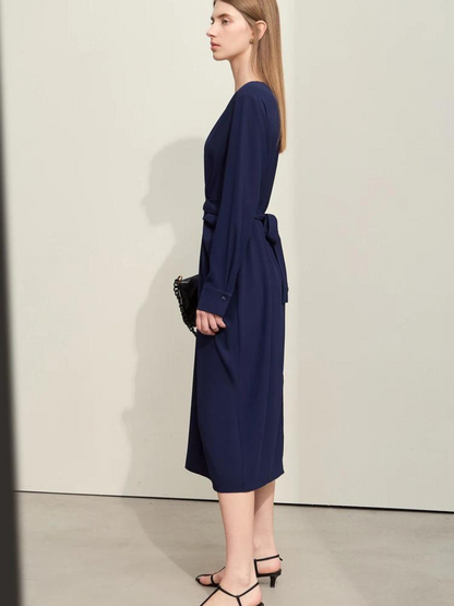 A woman wearing a dark blue, mid-calf length, long sleeved dress with a v-neckline and a draping design on the bodice and a bow on the back to cinch the waist.