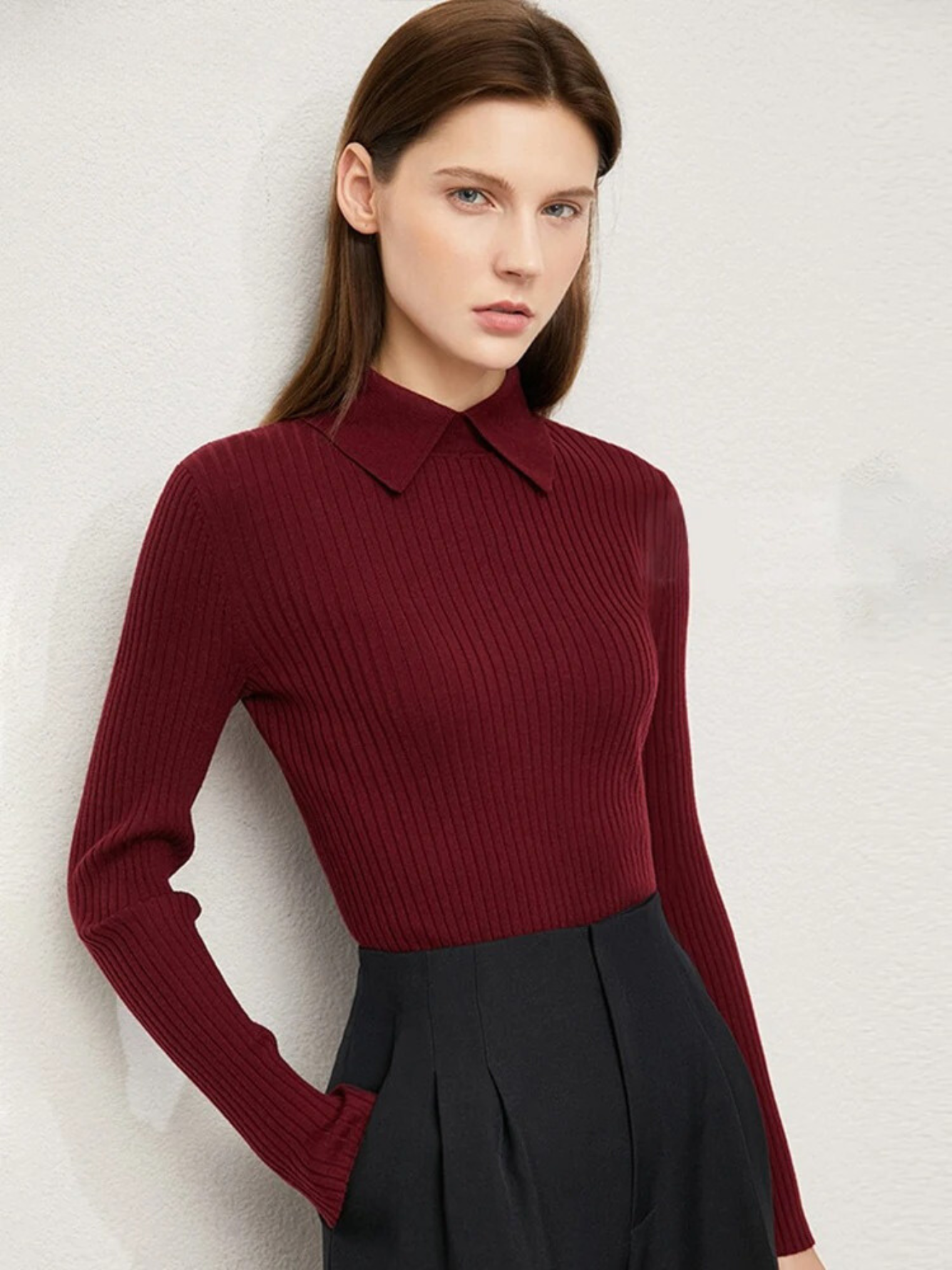 A brunette woman leaning against a white wall wearing a deep red sweater with a polo inspired collar and pleated black bottoms.