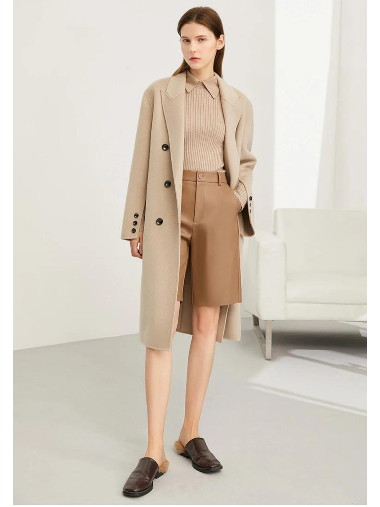 A brunette woman wearing a camel-colored sweater with a polo inspired collar, a pair of leather khaki shorts, a camel-coloured long coat, and leather mules.