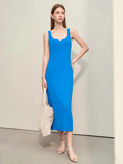 A woman wearing a minimalist, blue, mid-calf length dress with thin straps and a small cut out design on the neckline paired with simple open toed sandals and a bag.