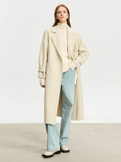 A woman wearing a cream colored knit turtleneck sweater with a tie design on both sides and a chevron knitting pattern, a cream colored oversized coat, and pair of light washed denim jeans.